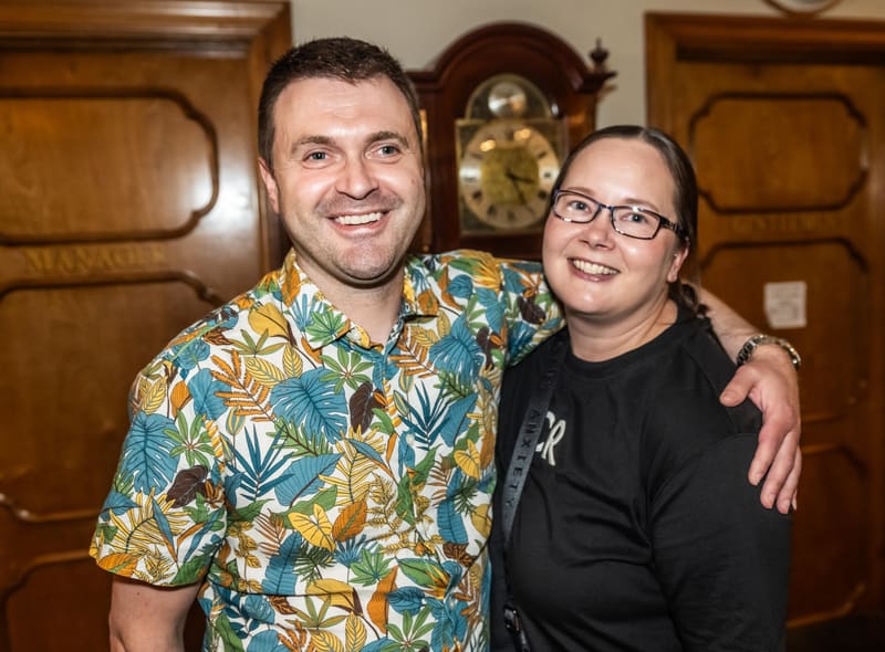 Two people smiling at the camera with their arms around each other in front of wooden panelling and a clock.