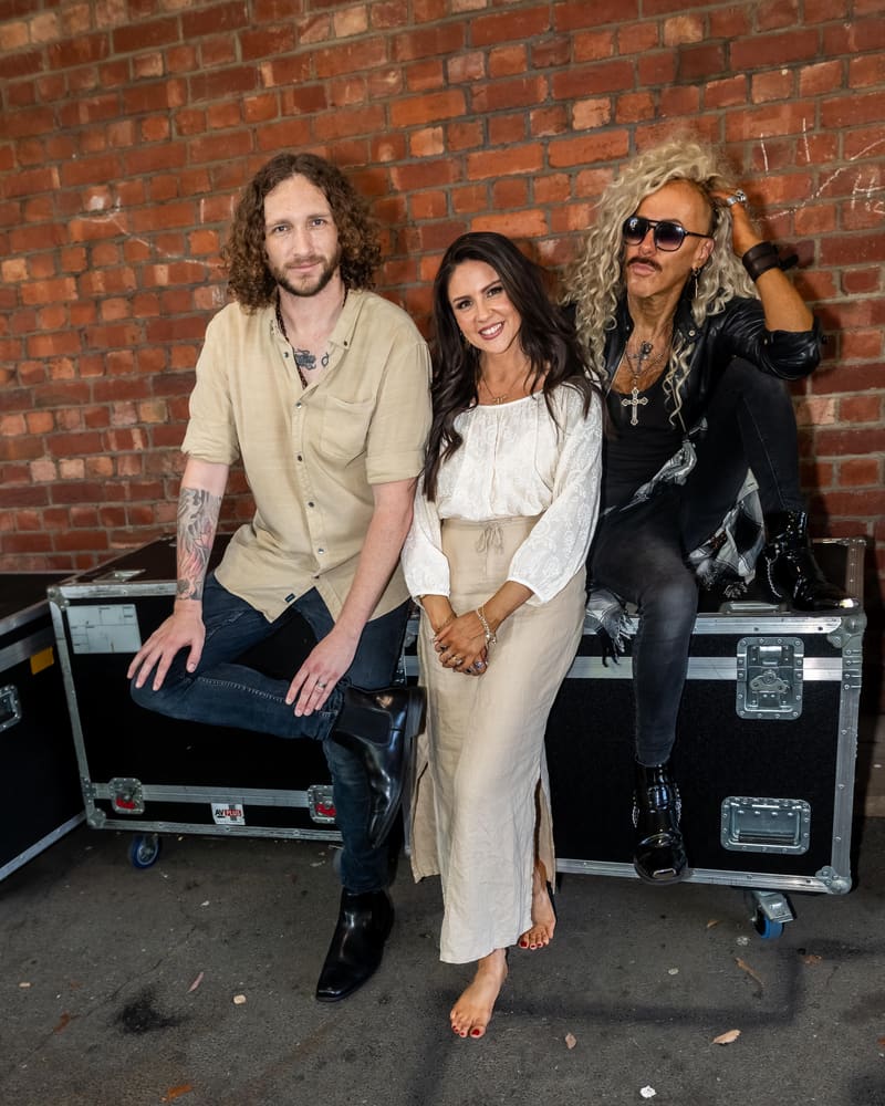 Three musicians pose together in front of a red brick wall and equipment cases.
