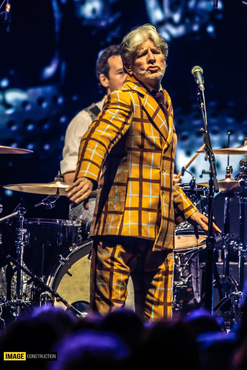 Performer in a bold yellow and black checked suit singing at a microphone on stage with a band and audience silhouettes visible.
