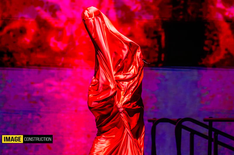 Performer in shimmering red costume with arms raised against vibrant magenta and purple stage lighting.