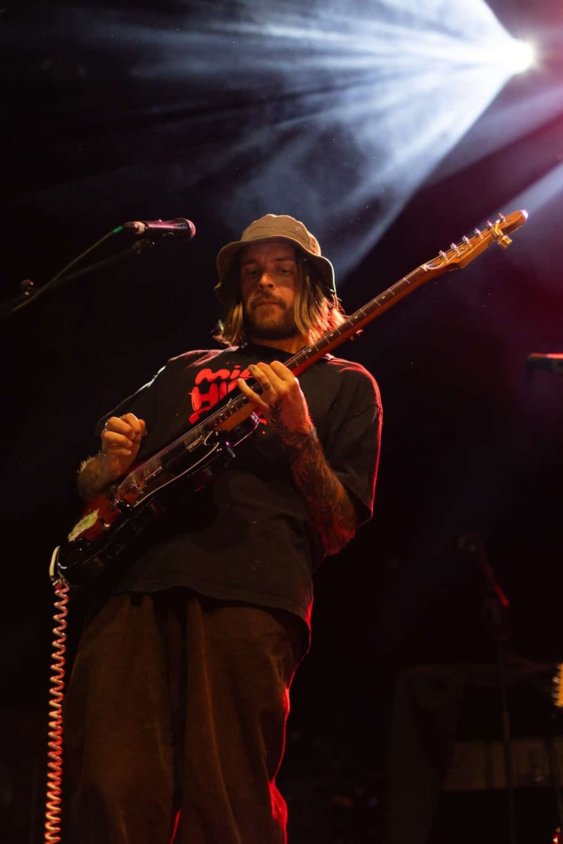 Guitarist wearing a hat and red shirt performing on stage under warm spotlight illumination.