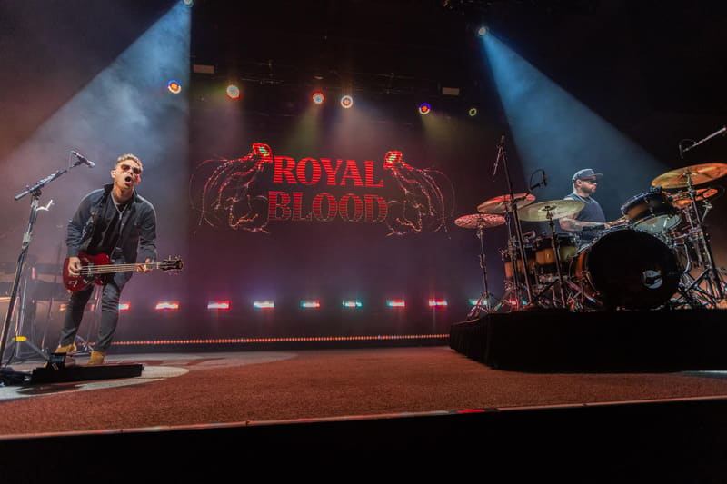 Royal Blood band performs on stage with guitarist and drummer beneath illuminated red logo and blue stage lighting.