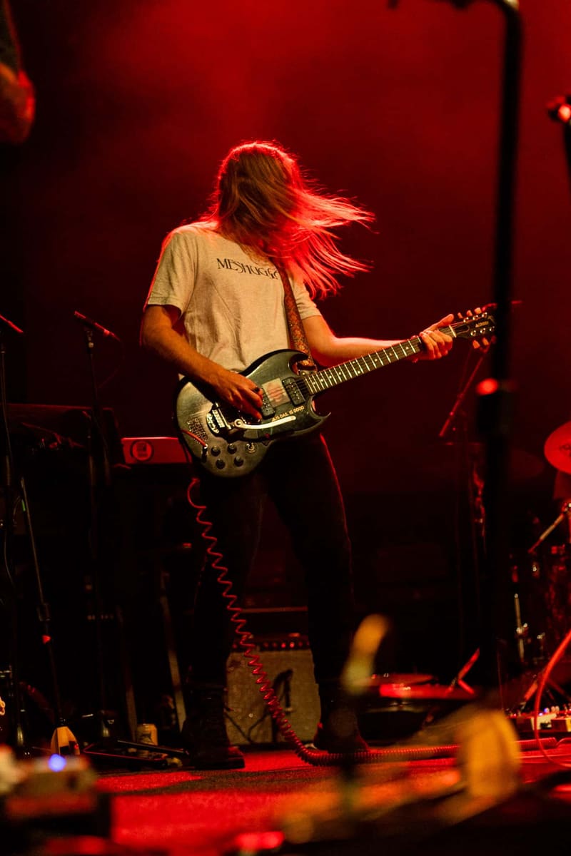 Guitarist with long hair playing electric guitar on stage under red lighting during a live performance.