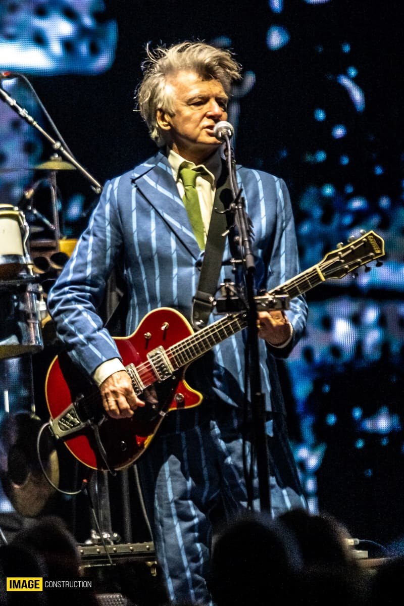 Musician performing with red electric guitar on stage, wearing a blue pinstriped jacket and yellow shirt beneath blue stage lighting.