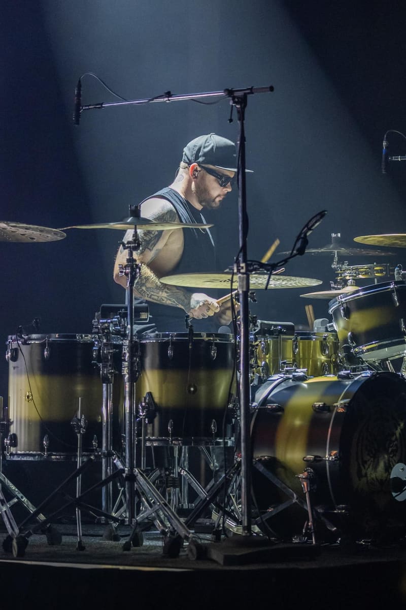 Drummer in a cap and sleeveless shirt performing at a drum kit under stage spotlights.