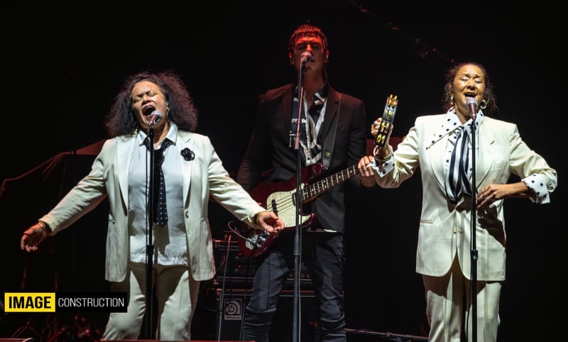 Three musicians perform on stage in light-coloured suits, with guitarist and saxophonist under warm stage lighting.