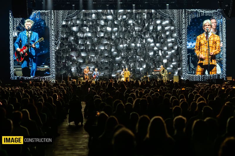Large crowd facing a stage with two giant screens displaying band members, central falling light effects, and yellow-lit performer visible centre stage.