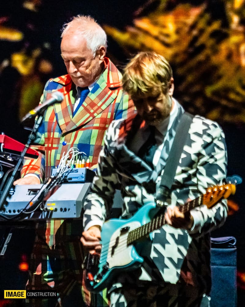 Two guitarists performing on stage, one in a colourful tartan jacket and one in a patterned dark jacket, under warm stage lighting.