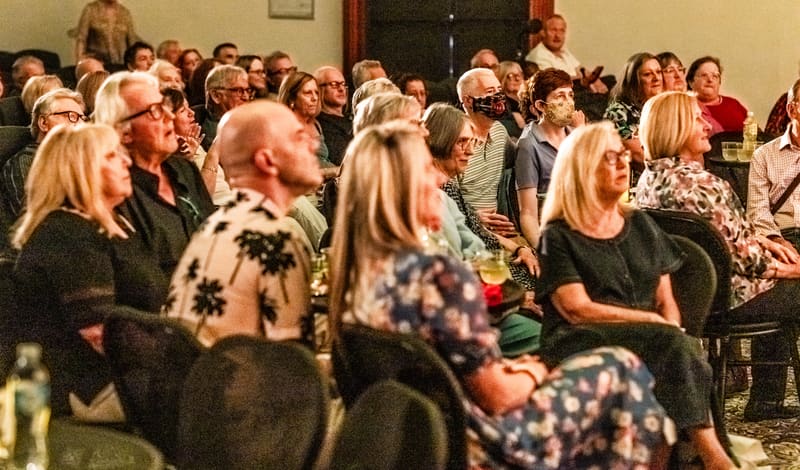 Seated gallery visitors watch an art event in an intimate indoor venue, with warm lighting illuminating the assembled crowd.