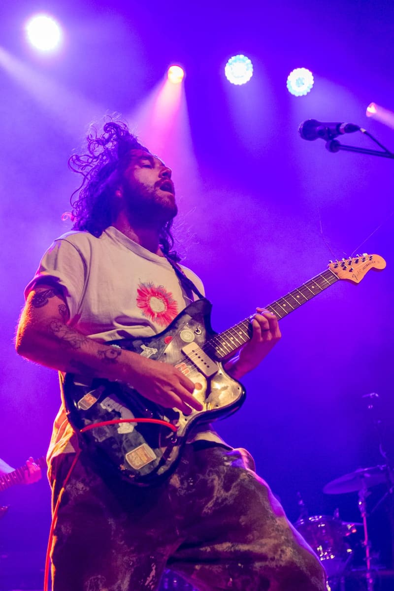 Guitarist performs on stage bathed in purple and pink stage lighting during a concert.