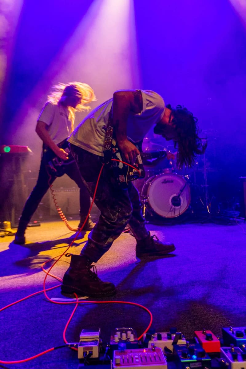 Band members perform on stage bathed in purple and yellow stage lighting, with drum kit visible in background.