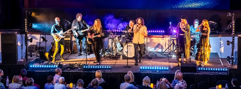 A band and ensemble performers in yellow and dark clothing play together on a lit stage before an audience.
