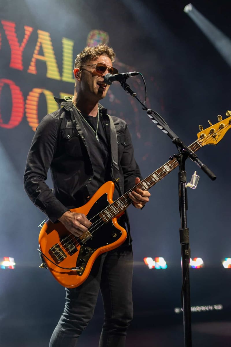 Musician performs on stage with orange bass guitar and microphone, wearing sunglasses and black jacket.