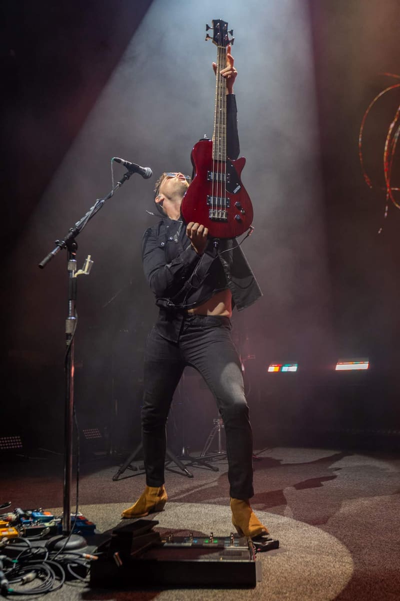 Bassist in black clothing and gold shoes performing with a deep red bass guitar under stage spotlights.