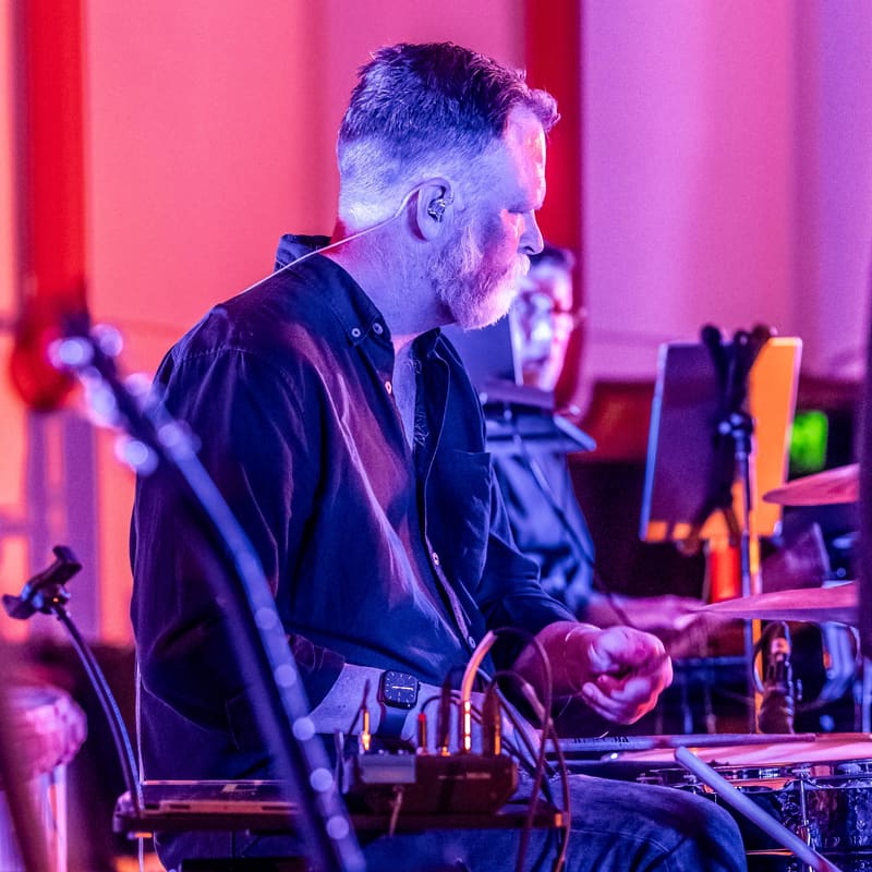 Musician performing at electronic equipment under red and purple stage lighting.