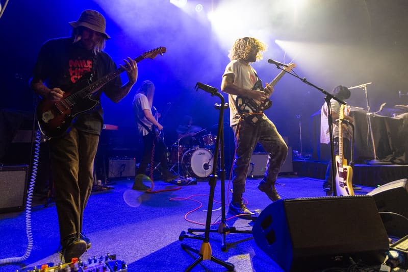 Band members perform on stage under blue and white spotlights, with guitarist in hat on left and blonde vocalist centre stage.
