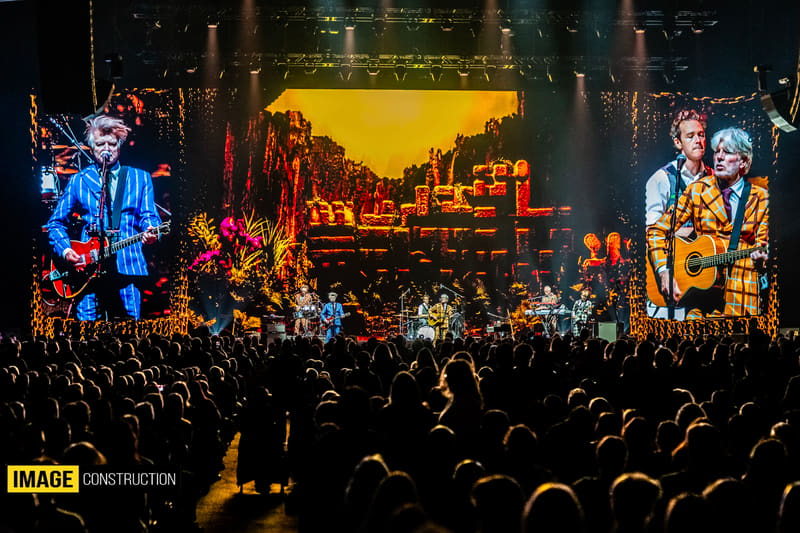 Band members perform on stage with giant video screens above a crowd of thousands during a concert tour.