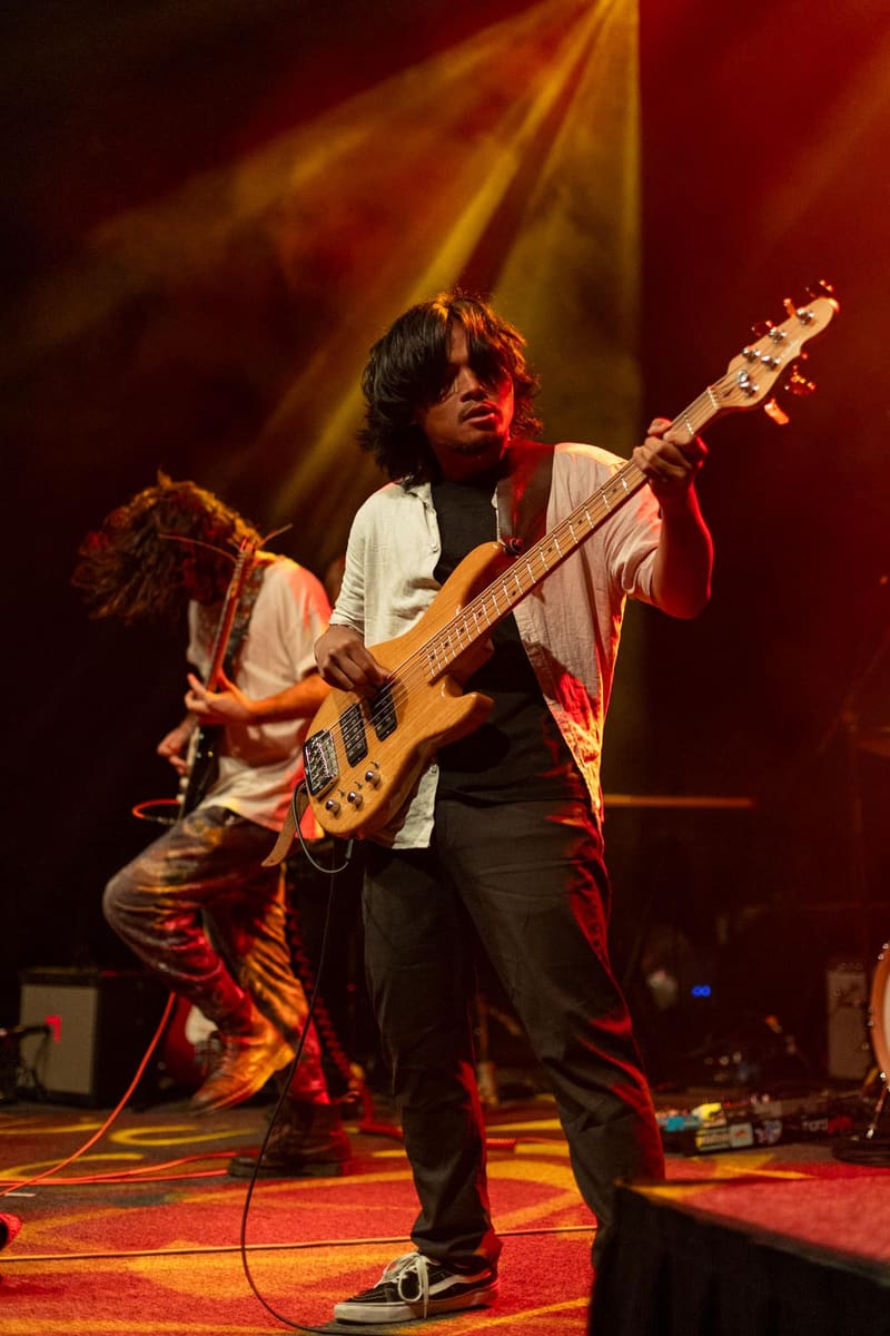 Guitarist performing with electric bass guitar under warm stage lighting, with band member visible in background.