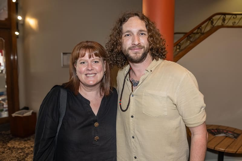 Two people posing together at a gallery opening, with warm lighting and wooden architectural details visible behind them.