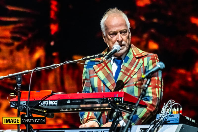 Musician performing at keyboard under warm stage lighting wearing a colourful patterned jacket.