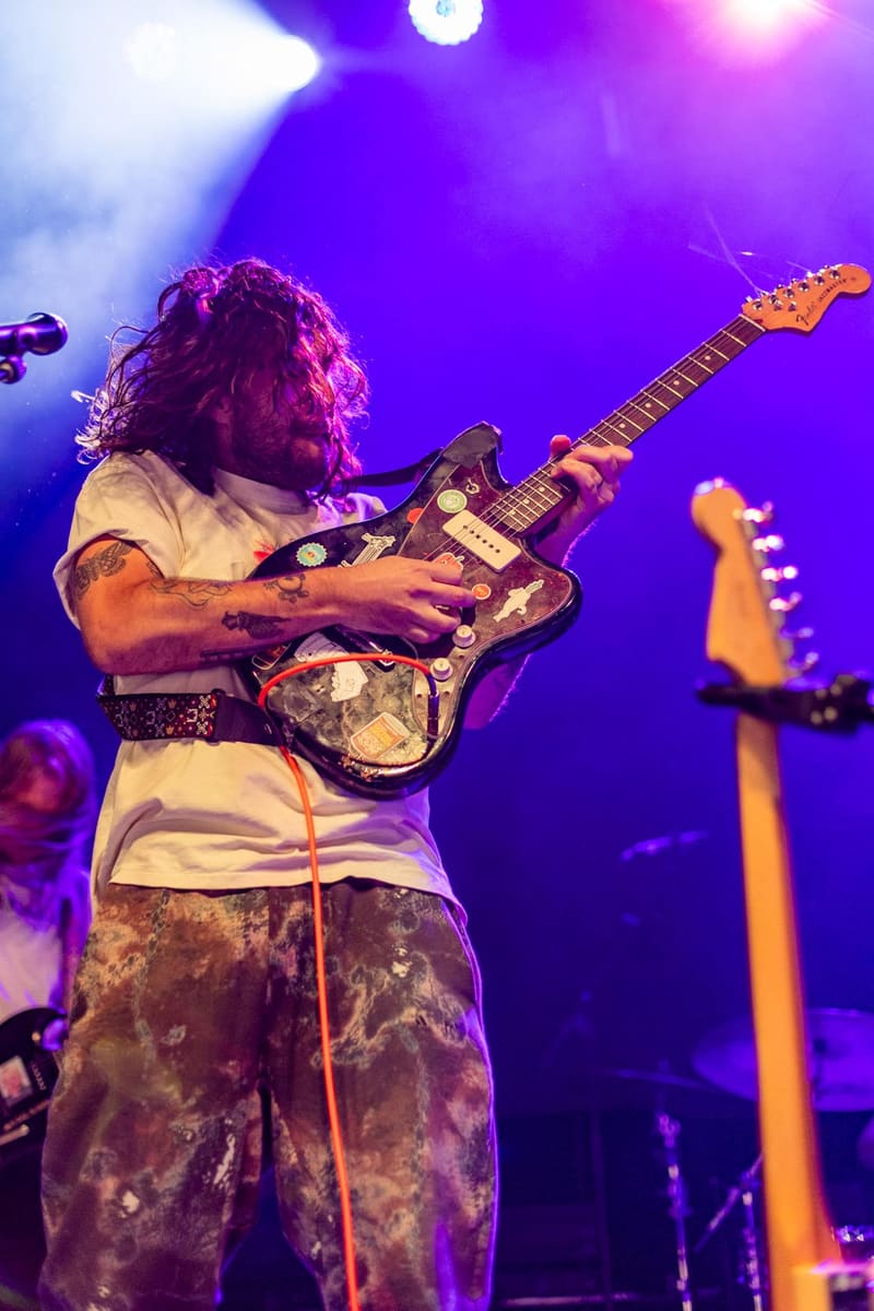 Guitarist with curly hair playing an electric guitar on stage under vibrant purple and blue lighting during a live performance.
