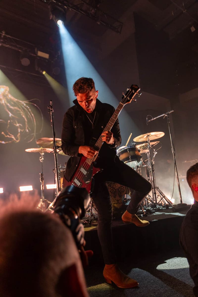 Bassist performing on stage under blue spotlight with drums and audience members visible.