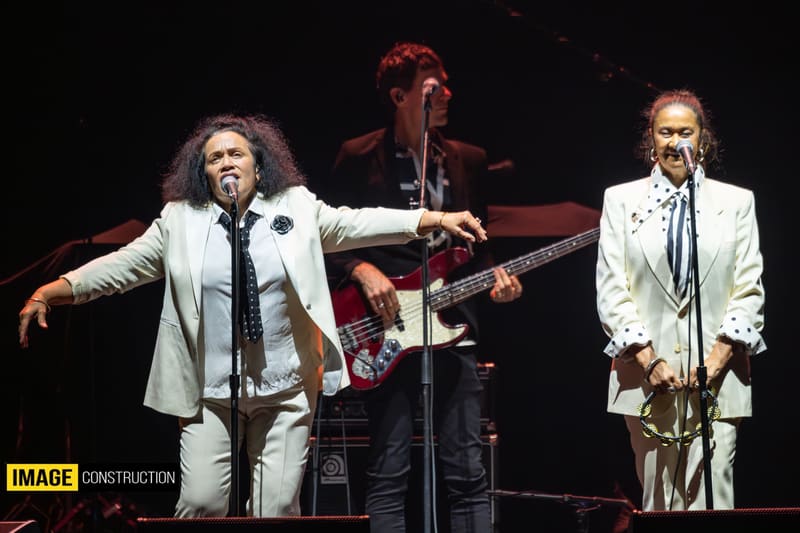 Three musicians perform on stage, two in white suits at microphones and one in dark clothing playing guitar, under warm stage lighting.