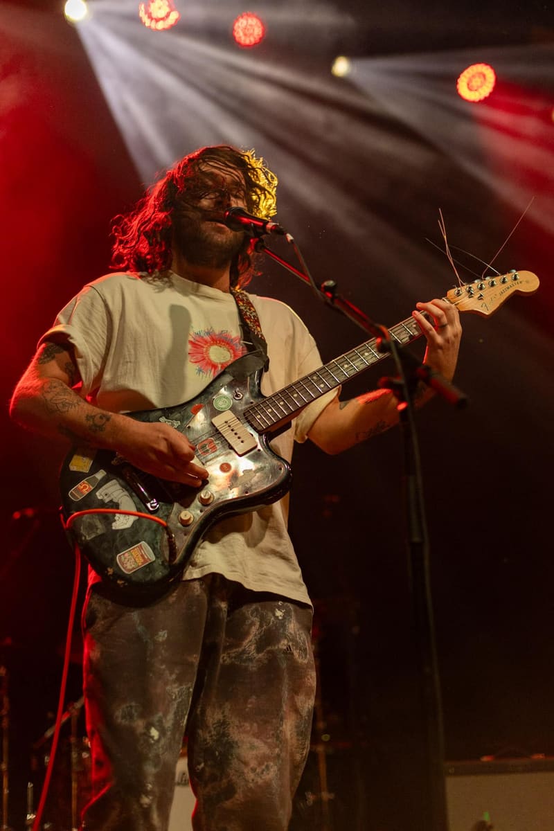 Guitarist with long hair and beard performing at a microphone under red stage lighting, holding a decorated electric guitar.