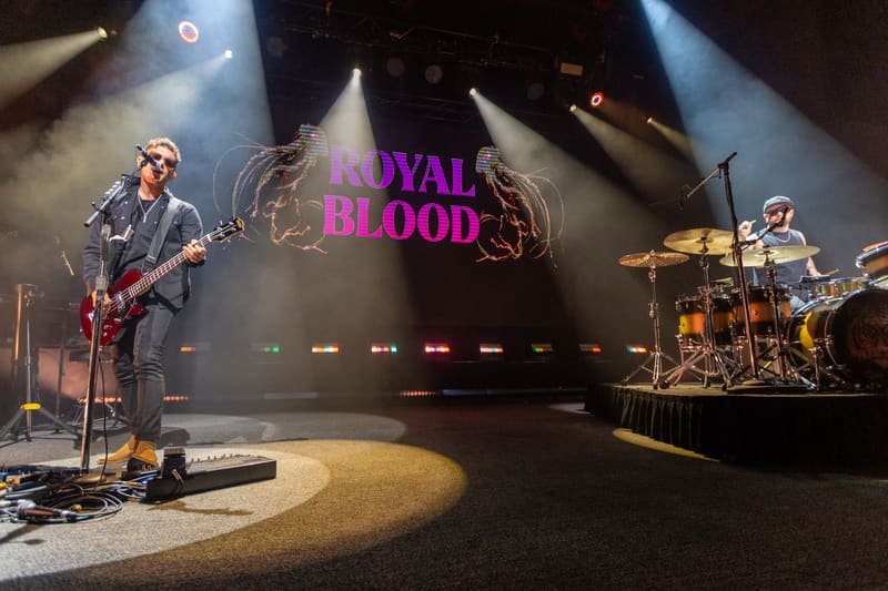 Royal Blood guitarist and drummer perform on stage with dramatic lighting and the band's magenta logo projected behind them.