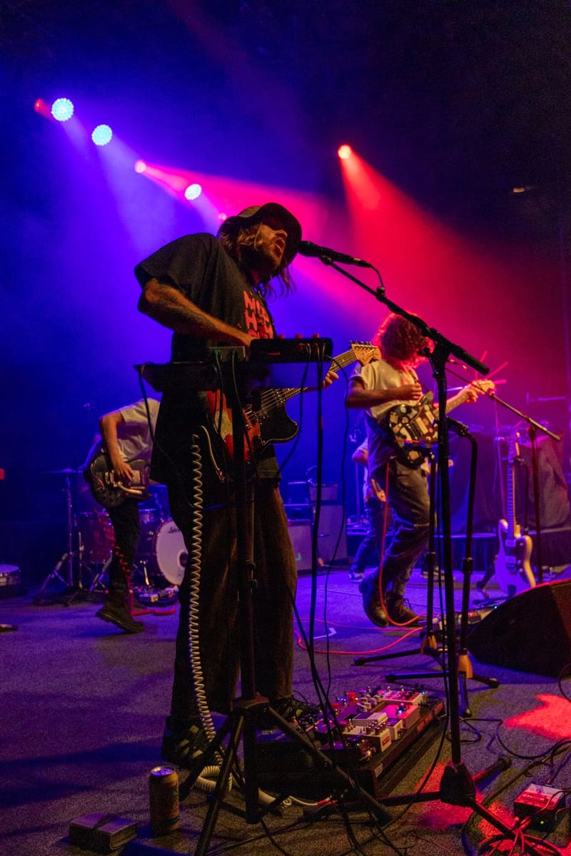 Musician with long hair and beard performs at microphone under vibrant pink and blue stage lighting.