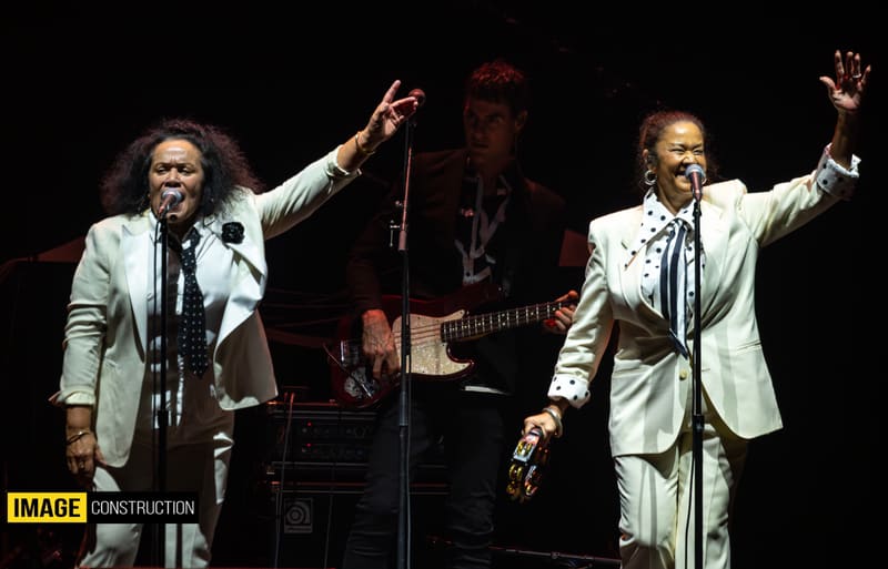 Two performers in cream blazers raise their arms on stage during the Forever Enz 2026 Tour, with a guitarist visible behind them.