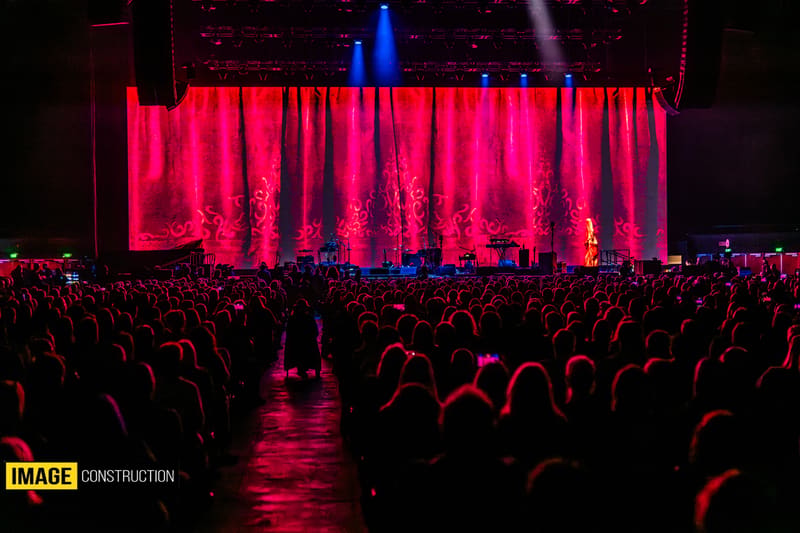 Large crowd faces a stage illuminated in pink and red light during Split Enz concert performance.