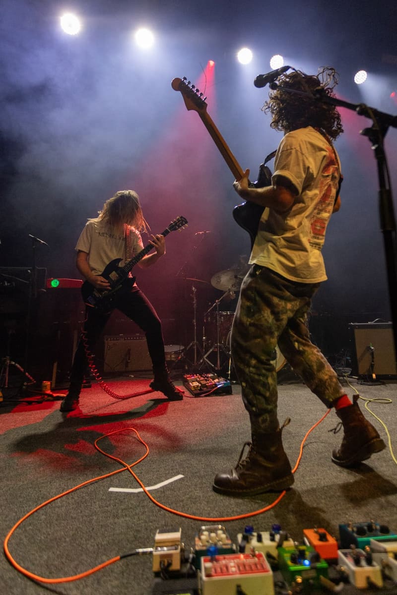 Two guitarists perform on stage under blue and pink spotlights, with effects pedals visible in the foreground.