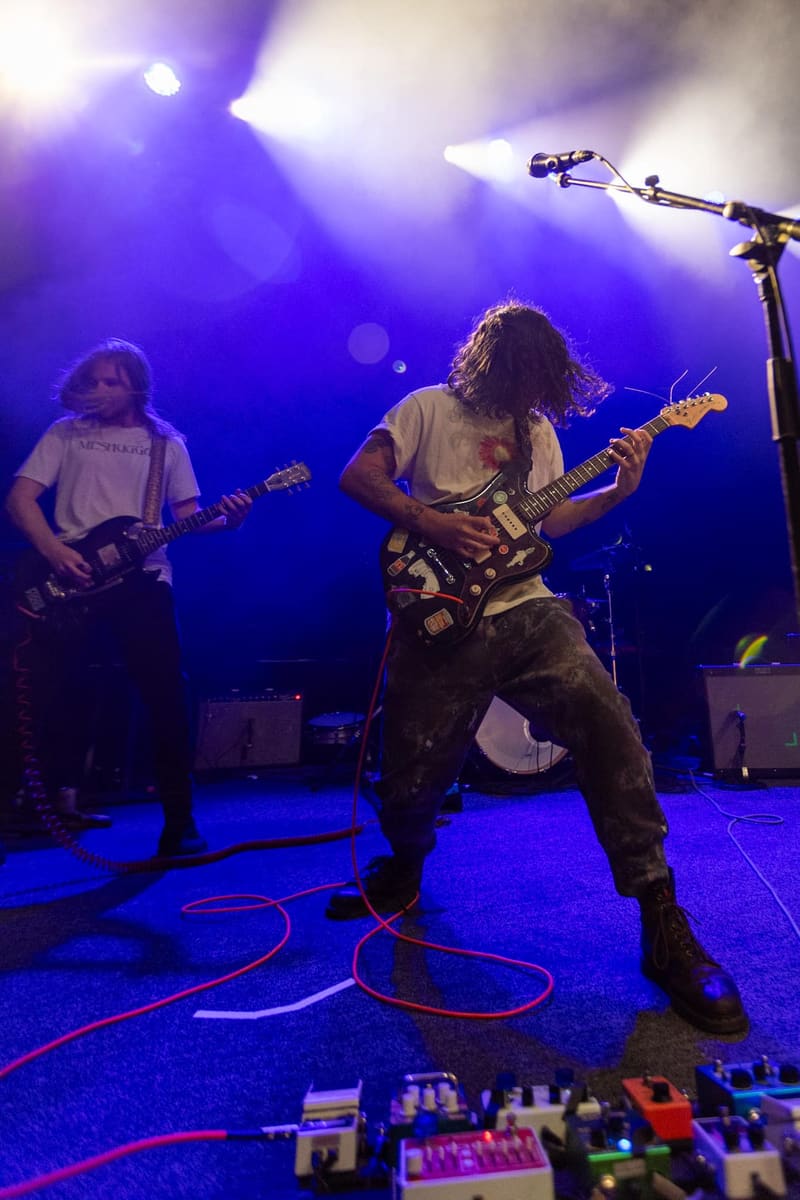 Two guitarists perform on stage under purple spotlights during a live rock performance.