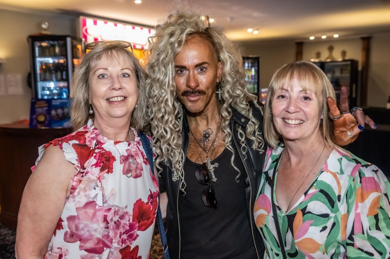 Three people pose together smiling at Gallery Three, with a man in the centre wearing long curly hair and dark clothing between two women in patterned outfits.