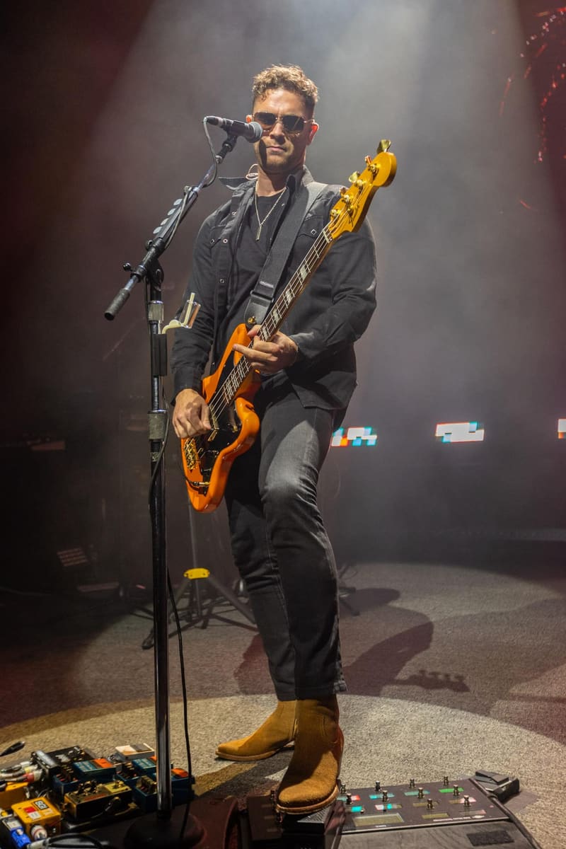 Guitarist in black jacket and sunglasses performs with an amber-coloured electric guitar under stage spotlights.