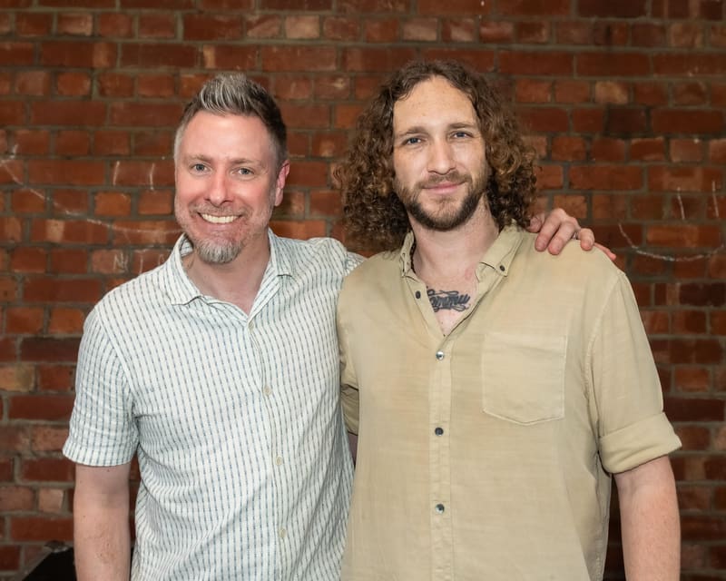 Two men stand side-by-side smiling at the camera against a red brick wall.
