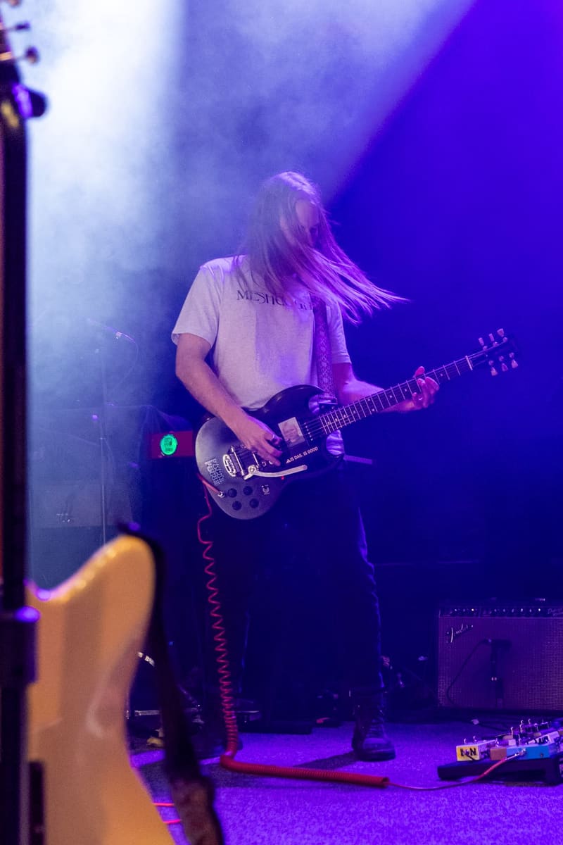 Guitarist with long dark hair performs on stage under purple lighting during a Psychedelic Porn Crumpets concert.