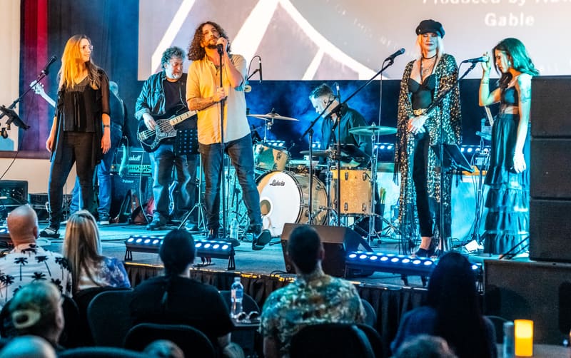 Five musicians perform on a stage bathed in blue and red lighting, with audience members visible in the foreground.