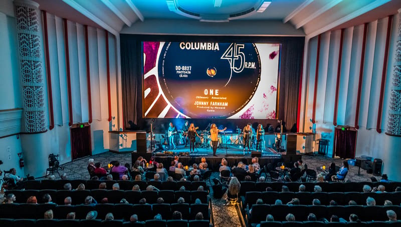 Musicians perform on stage beneath a large projection screen in a theatre auditorium filled with seated audience members.
