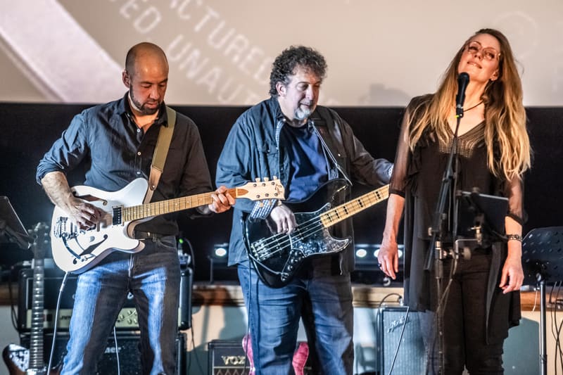 Three musicians perform on stage: two guitarists in denim shirts and a female vocalist in black, under warm stage lighting.