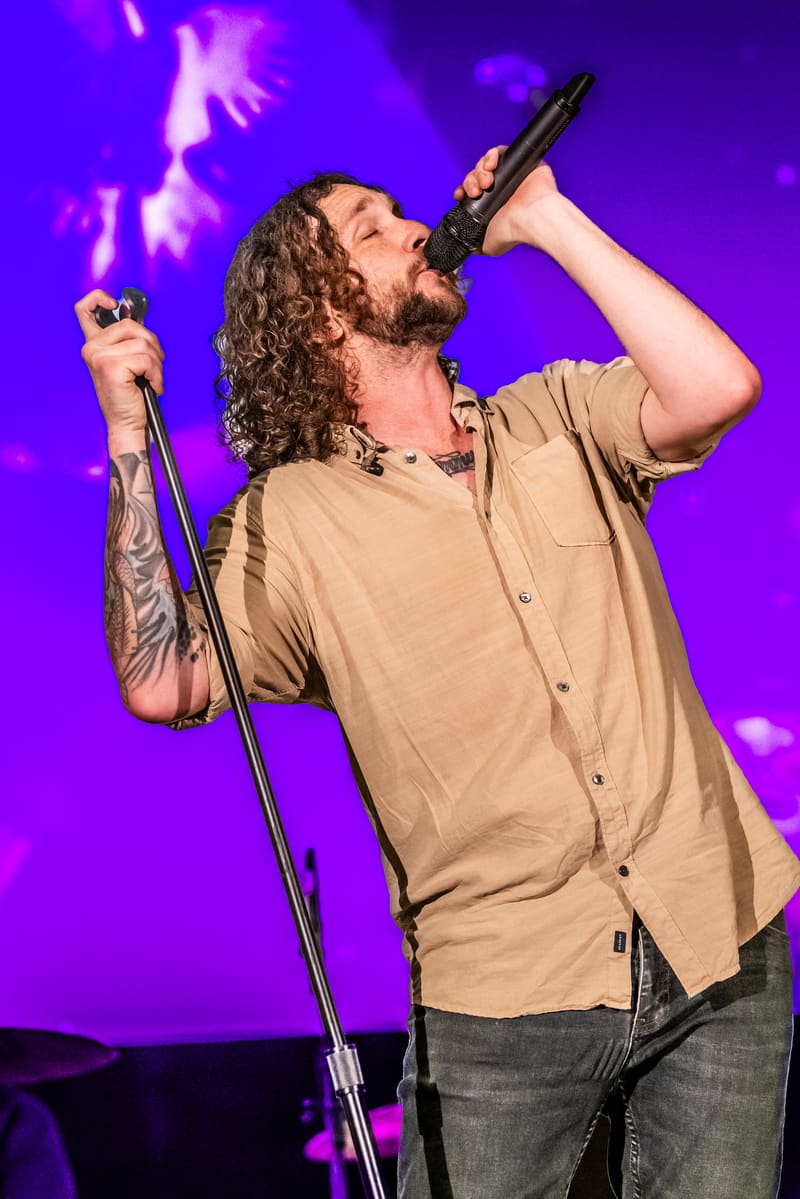 Singer with long curly hair performing with dual microphones against purple stage lighting and atmospheric effects.