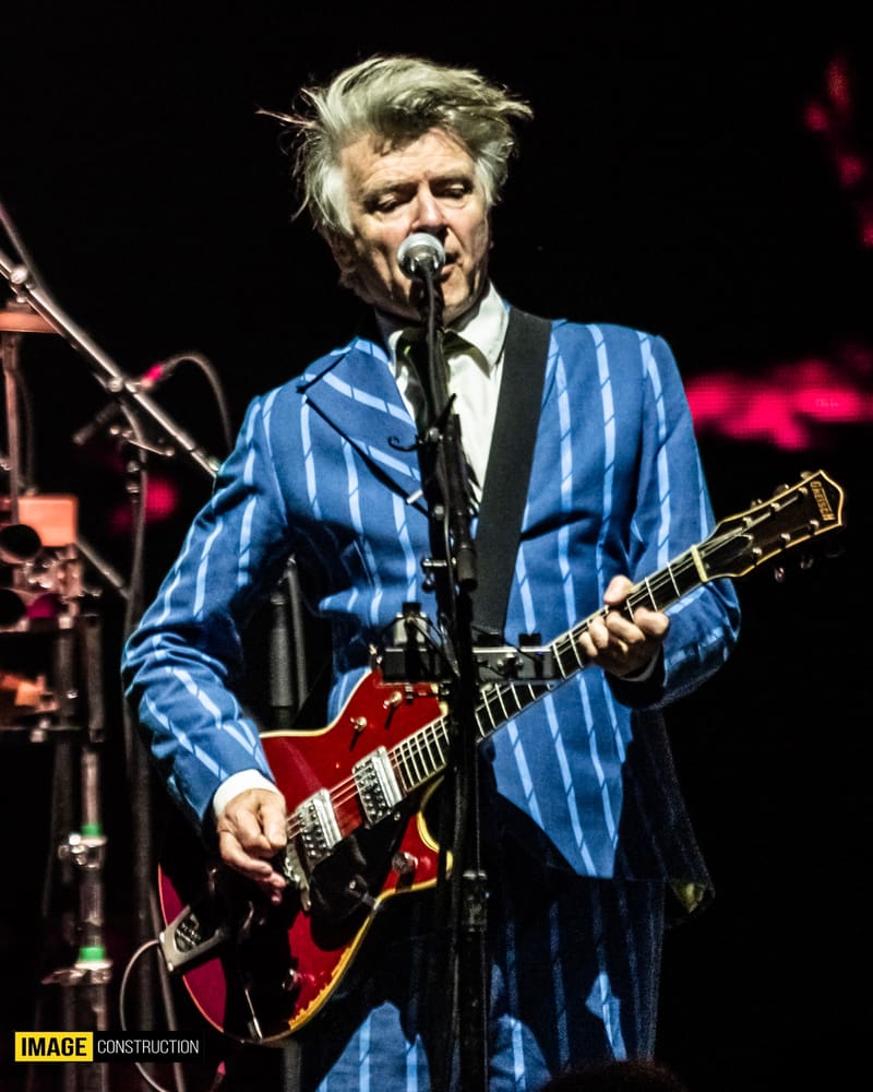 Musician in blue pinstriped jacket playing red guitar at microphone on stage with warm lighting.