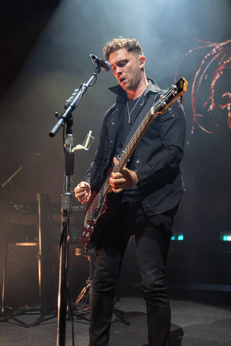 Guitarist and vocalist performing at a microphone in black clothing against a moody stage backdrop.