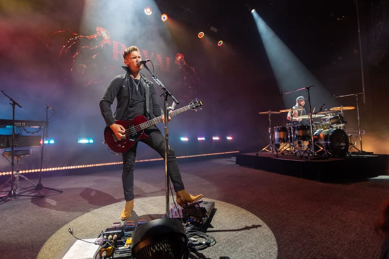 Guitarist performing on stage with drummer visible in background, lit by atmospheric blue and warm spotlights.