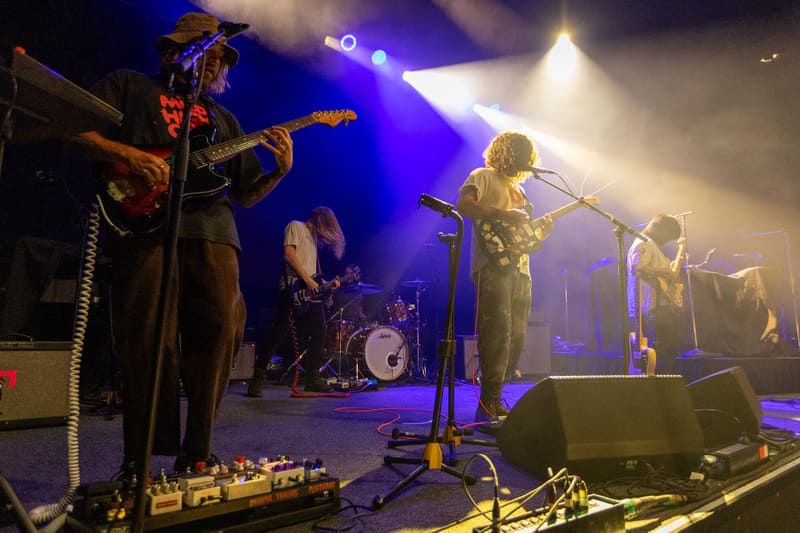 Psychedelic Porn Crumpets perform on stage under blue and white spotlights with guitarist, drummer, and vocalist in silhouette.