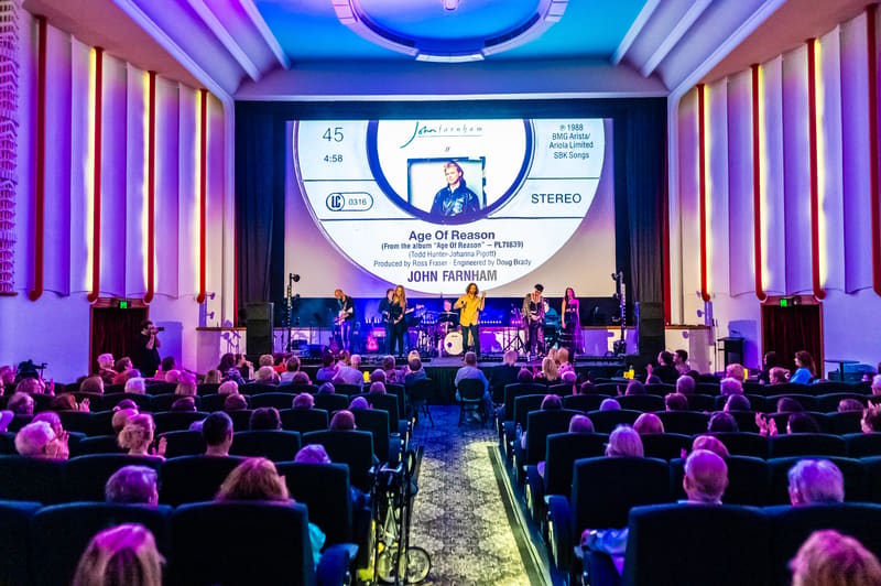 Large audience seated in purple-lit theatre facing a stage with band performing beneath a vintage record sleeve projection.