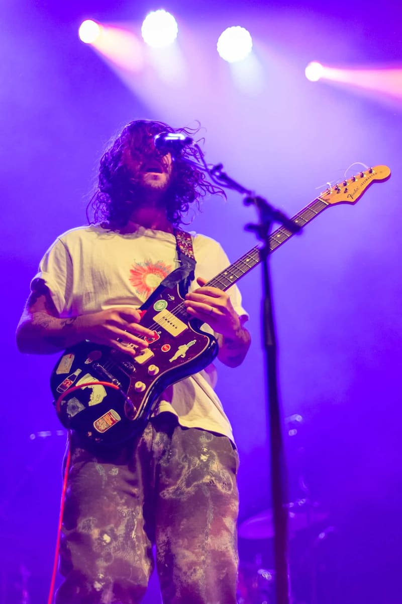 Guitarist with long dark hair performs under purple stage lighting, wearing a graphic t-shirt and patterned pants.