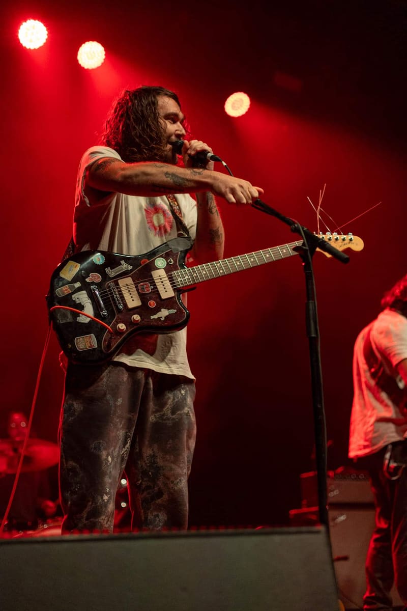 Musician with long dark hair singing into a microphone while playing an ornate black electric guitar, bathed in red stage lighting.
