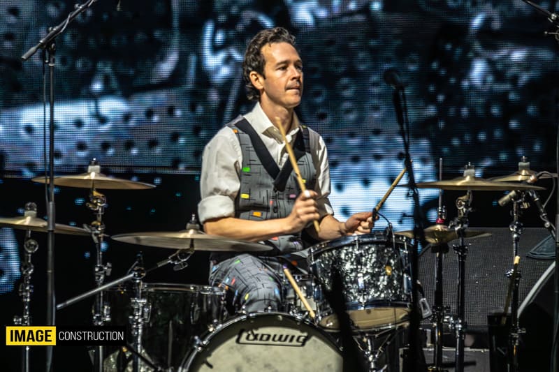 Drummer performing at kit with microphone in front of illuminated stage backdrop during Split Enz tour.